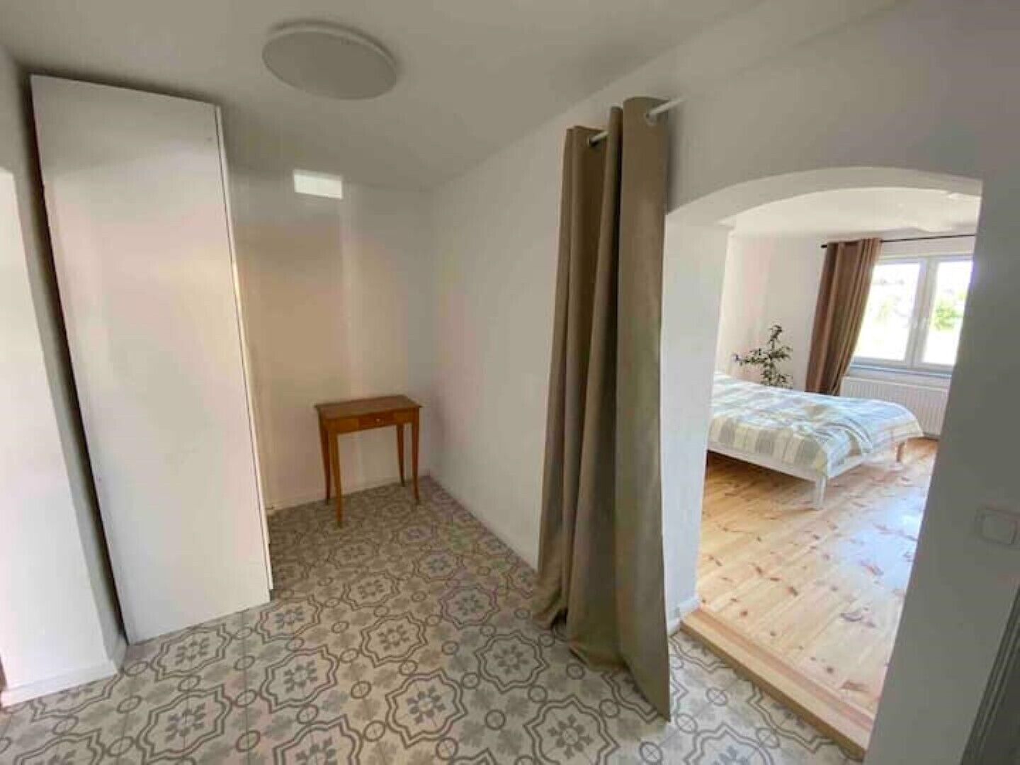 A room with a patterned floor, a wooden table, a bed with a patterned bedspread, and a window with curtains.
