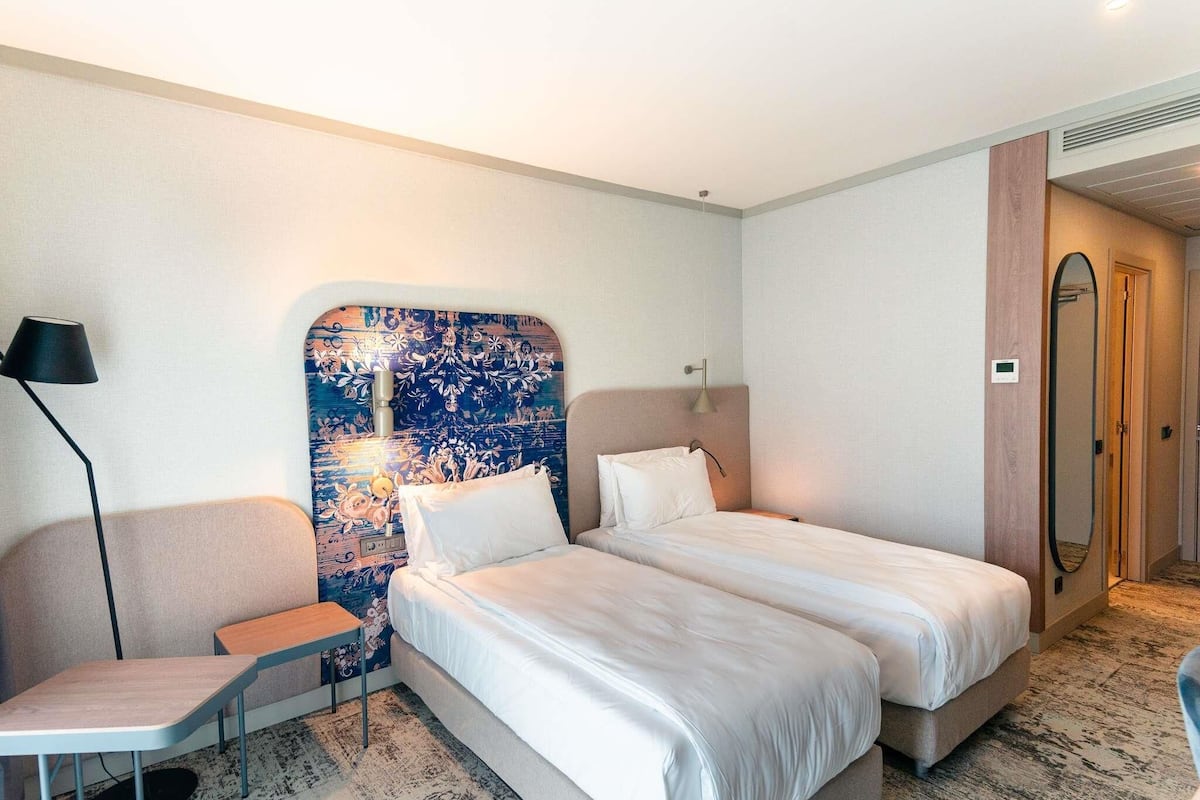 Ein Hotelzimmer mit zwei Betten, einem Schreibtisch, einem Stuhl, einem groĂen Spiegel und einem dekorativen Kopfteil.