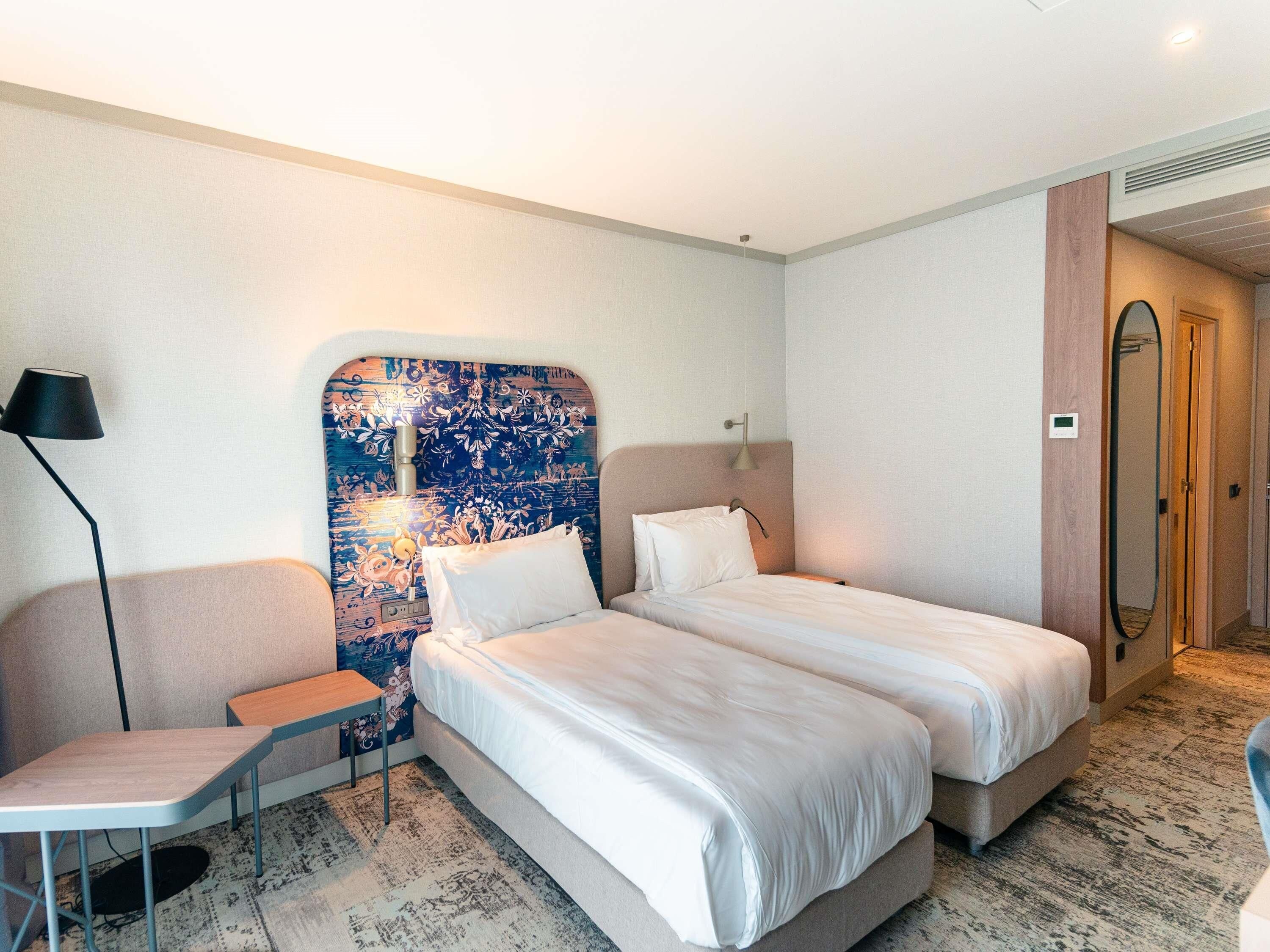 A hotel room with two beds, a desk, a chair, a large mirror, and a patterned headboard.