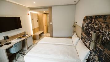 A hotel room with a bed, a desk with a chair, a TV, and a bathroom visible through an open door.