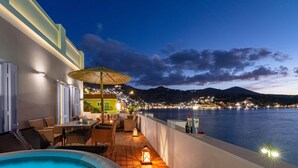 Une terrasse extérieure avec une piscine, une plateforme en bois, des meubles en osier et une vue sur une ville côtière la nuit.