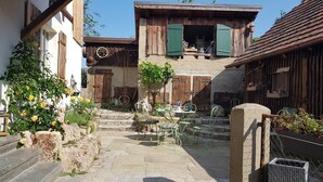 A courtyard with stone steps leading to a wooden building with green shutters. There are outdoor tables and chairs, and a small garden with yellow flowers. - Romantic studio in renovated half-timbered house from 1706, with courtyard and garden (Bodman-Ludwigshafen)
