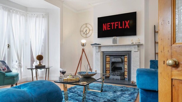 A living room with a fireplace, a television displaying 'NETFLIX', and a blue sofa. - Huge House Pool Table Parking Garden sky tv Wifi (Gloucester)