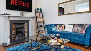 A living room with a fireplace, a blue sofa with decorative pillows, a glass coffee table with a fruit and cheese board, and a TV mounted above the fireplace displaying the Netflix logo. - Huge House Pool Table Parking Garden sky tv Wifi (Gloucester)