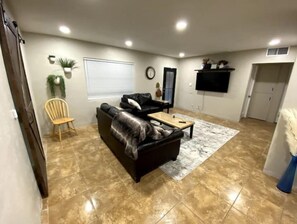 Relax in this stylish living area with a sofa, armchair, and coffee table, featuring a wall-mounted TV and large window with blinds.
