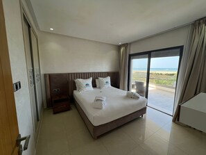 A hotel room with a large bed, a nightstand, a desk, and a view of the beach.