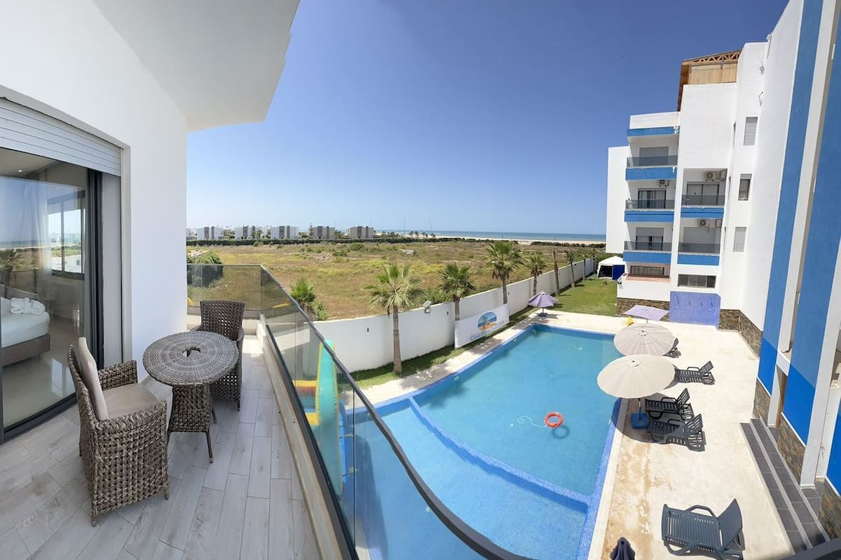 A balcony with a bed, a round table, and wicker chairs. There is a swimming pool with lounge chairs and umbrellas.