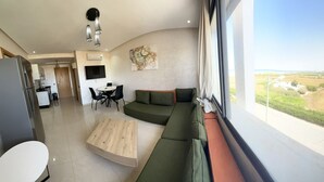 Un salon moderne avec un canapé aux lignes courbes, une table basse en bois et un coin repas comprenant une petite table et des chaises. Une grande fenêtre offre une vue sur un paysage rural.