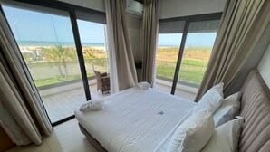 Une chambre d’hôtel dotée d’un grand lit, de linge de lit blanc et offrant une vue sur la plage et la végétation grâce à de grandes fenêtres du sol au plafond.