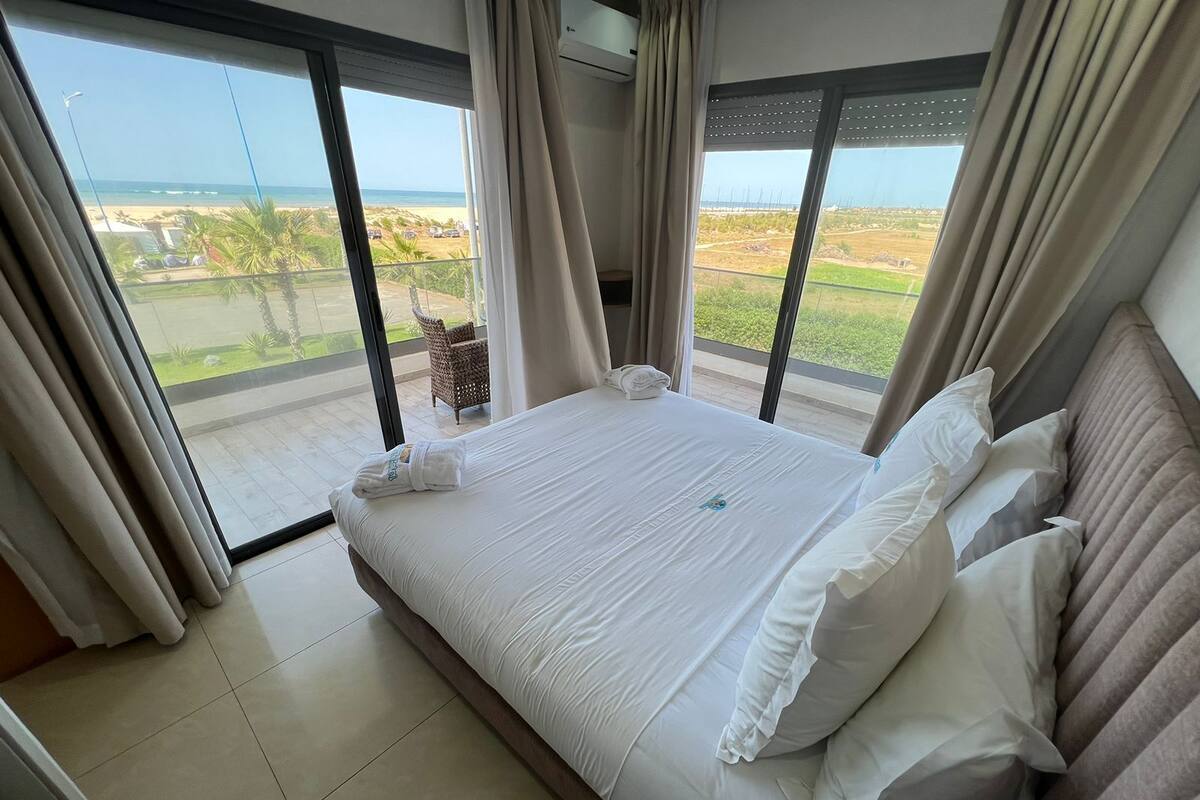 A hotel room with a large bed, white linens, and a view of the beach and greenery through floor-to-ceiling windows.