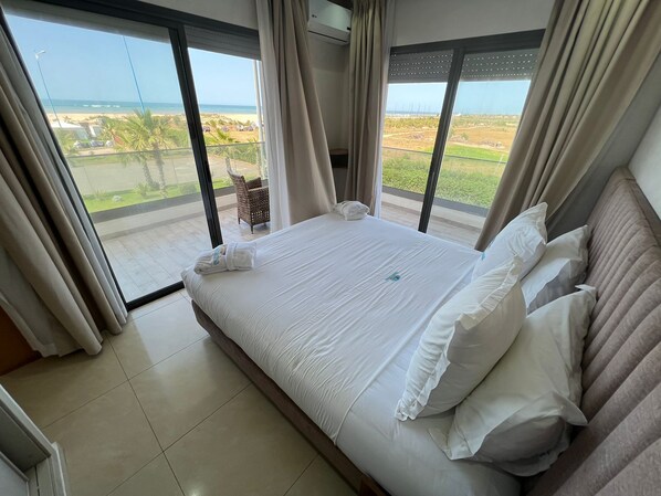 A hotel room with a large bed, white linens, and a view of the beach and greenery through floor-to-ceiling windows.