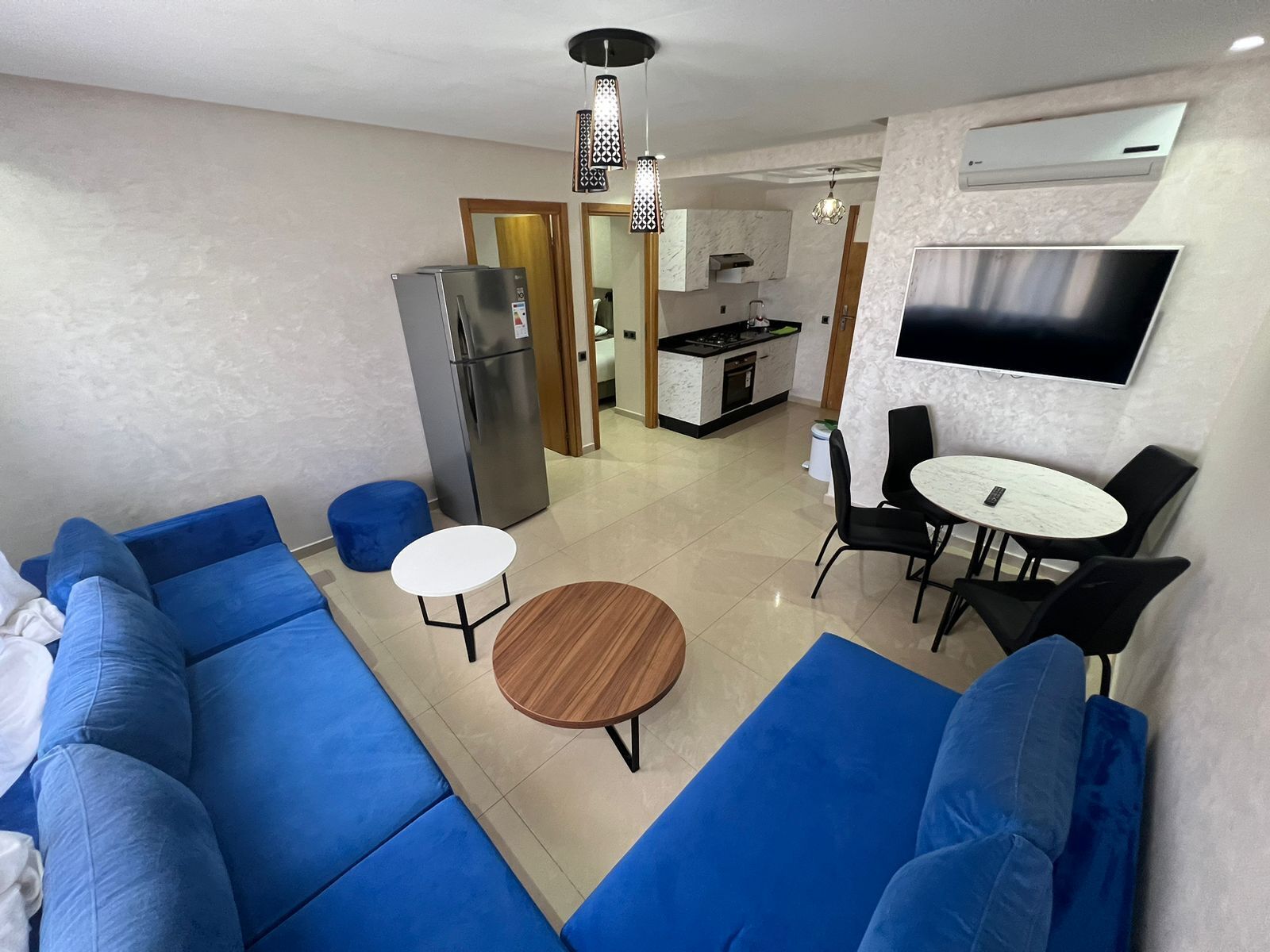 Uma sala de estar moderna com sofás azuis, uma mesa redonda de madeira e uma TV fixada na parede.