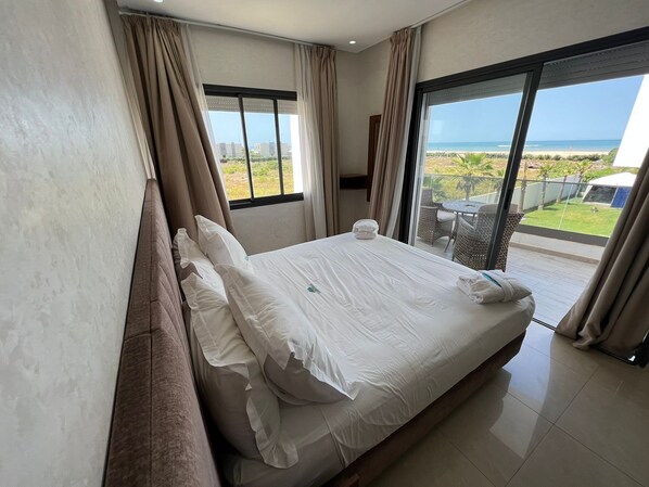 A hotel room with a bed, large windows, and a view of the beach.