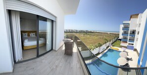Un balcón con una cama, una mesita y una zona de piscina con sombrillas y sillones de descanso.