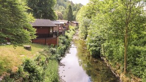 A row of wooden cabins with a river running between them, surrounded by lush greenery. - Nutkins Lodge (Windermere)
