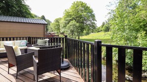 A wooden deck with wicker furniture and a glass table, overlooking a river and greenery. - Nutkins Lodge (Windermere)