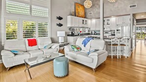 A spacious living area with a sofa, coffee table, and ottoman, a kitchen with white cabinets, and large windows with blinds. - Just Like Heaven - Beachfront (Crawfordville)