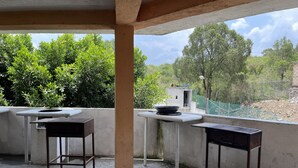 Un espacio exterior techado con pilares de concreto, una cerca metálica y vistas a árboles y un edificio lejano.