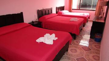 Habitación de hotel con tres camas, cada una con una colcha roja y toallas blancas. La habitación tiene baldosas de suelo estampadas y una ventana con cortinas.