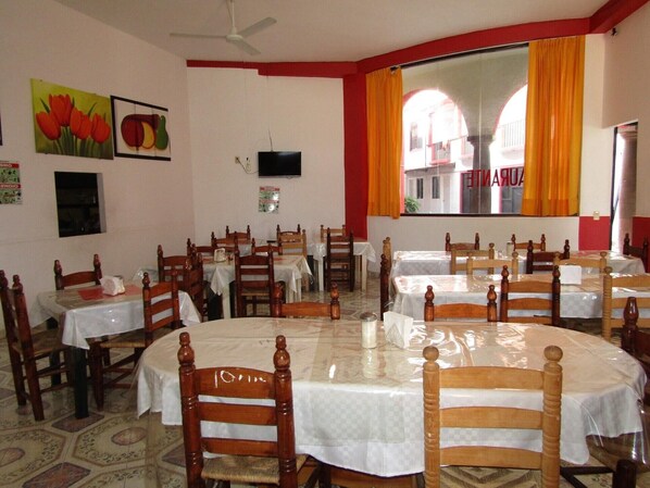 A dining area with round tables covered with white tablecloths, wooden chairs, and a television mounted on the wall.
