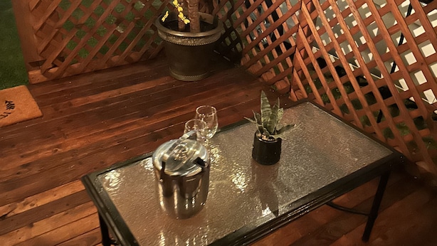 Una terraza de madera con una mesa de vidrio, una tetera de metal y plantas en macetas.