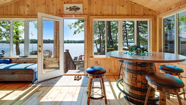 A wooden cabin with a large open door leading to a deck, a round bar table, and a bed with a view of the lake. - Lakefront Home with Sandy Beach! Kayaks, SUP, Pedal boat, and more! (Shapleigh)