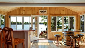 A wooden cabin with a bar area, large windows, and a view of the lake. - Lakefront Home with Sandy Beach! Kayaks, SUP, Pedal boat, and more! (Shapleigh)