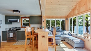 A spacious kitchen with wooden cabinets, a dining area with a table and chairs, and a cozy seating area with a sofa and large windows overlooking a body of water. - Lakefront Home with Sandy Beach! Kayaks, SUP, Pedal boat, and more! (Shapleigh)