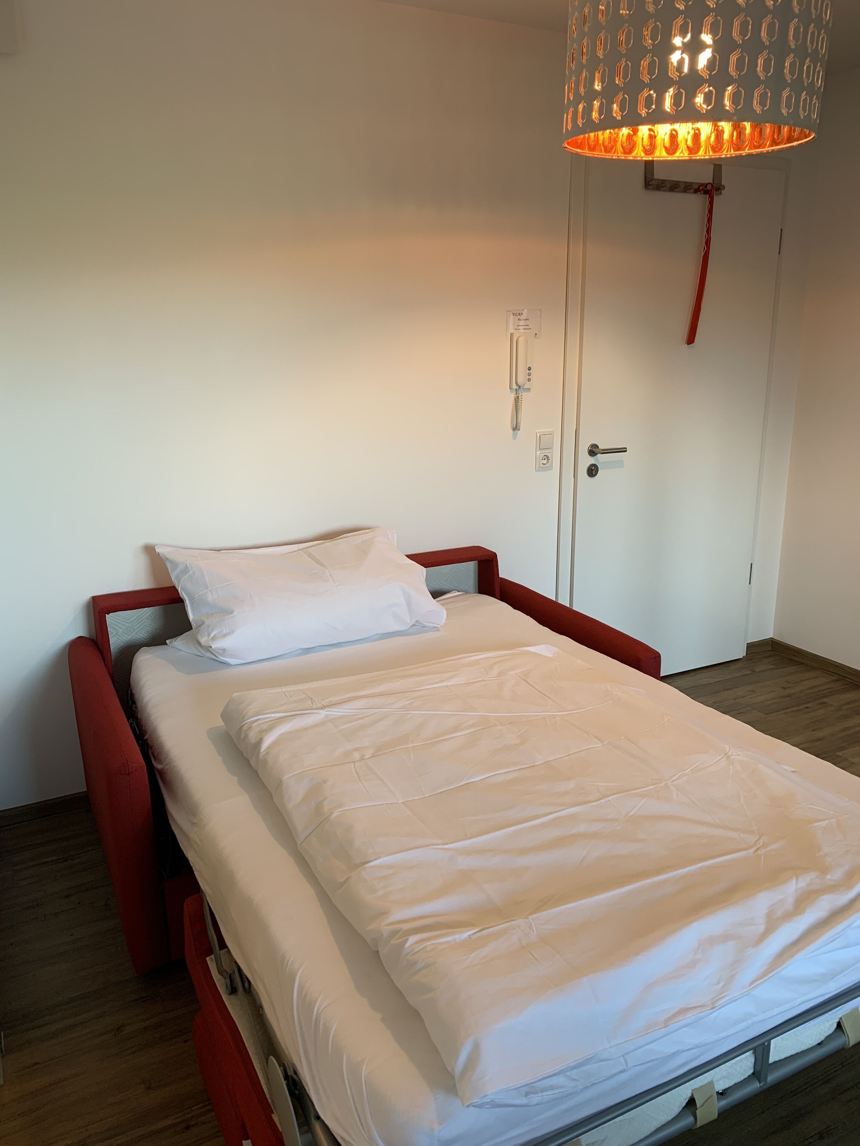 Un lit simple avec un matelas et des oreillers blancs, une tête de lit rouge et un luminaire suspendu au-dessus.