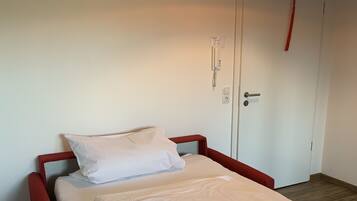 Un lit simple avec un matelas et des oreillers blancs, une tête de lit rouge et un luminaire suspendu au-dessus.