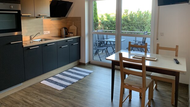 A modern kitchen with dark cabinets, a stainless steel sink, and a coffee maker. There is a dining area with a table and chairs, and a large window offering a view of greenery outside. - Vacation apartments Gergert (Löwenstein)