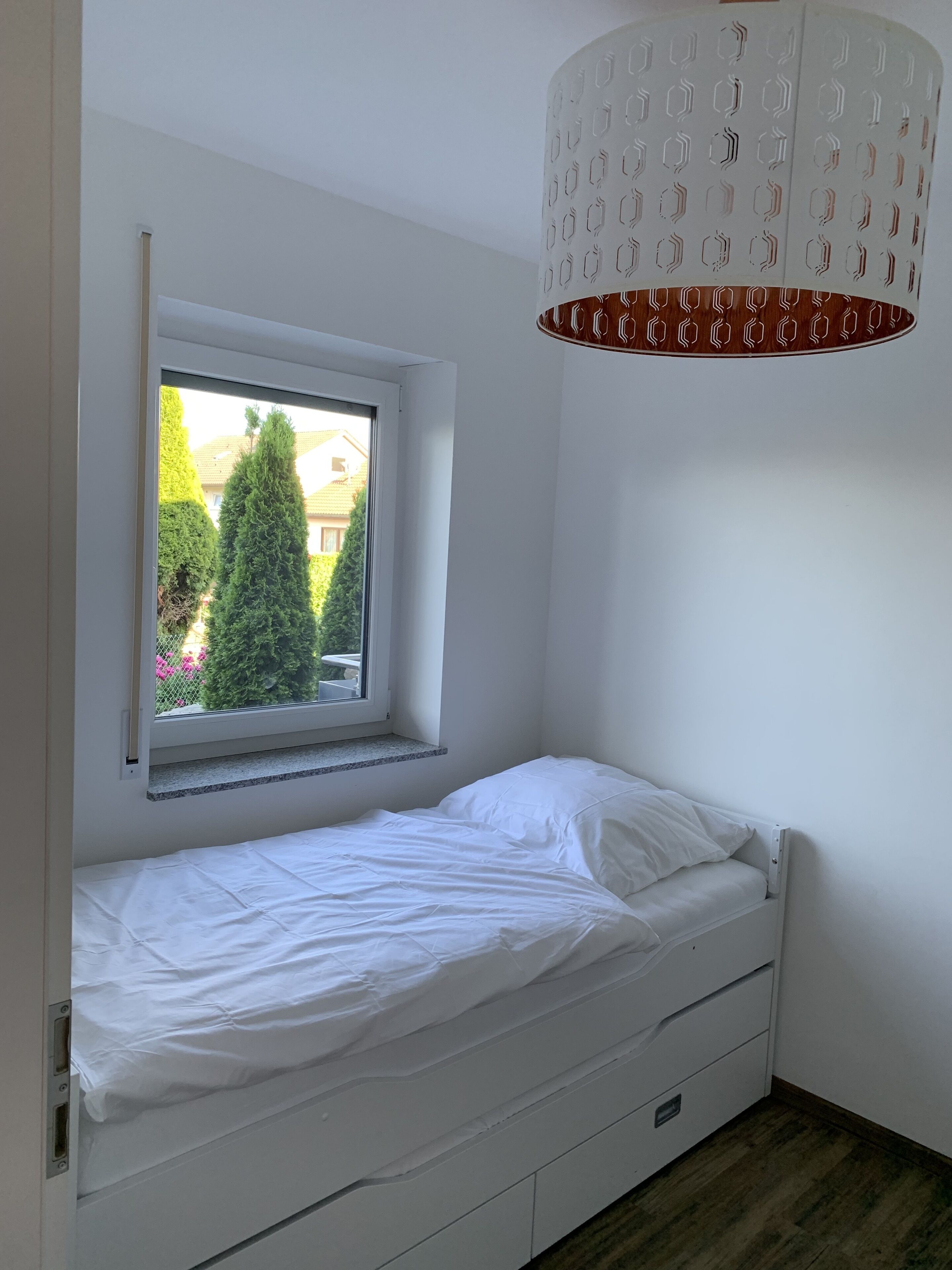 A small, white bedroom with a single bed, a window, and a ceiling light fixture.