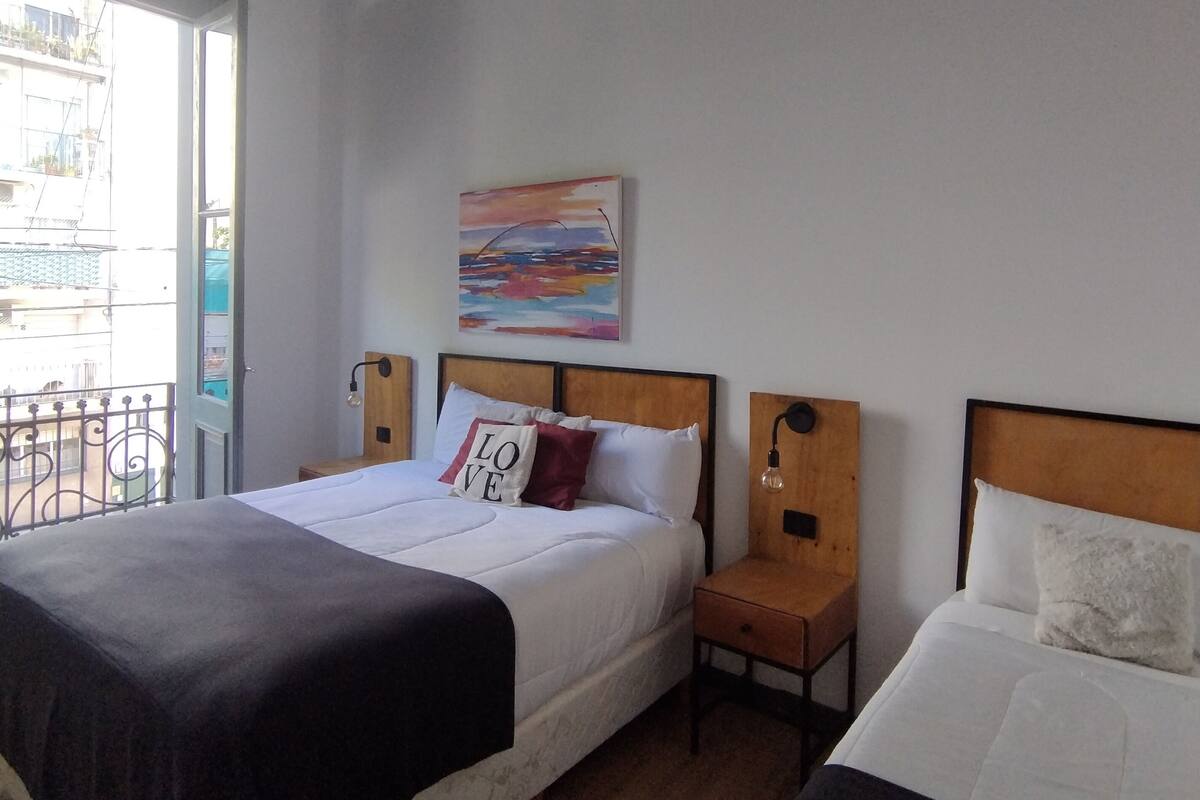 A hotel room with a large bed, a smaller bed, a nightstand with a lamp, a painting on the wall, and a balcony with a railing.