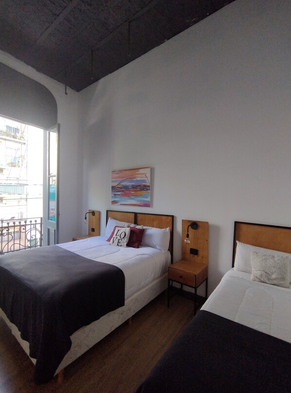 Habitación de hotel con una cama grande, una cama más pequeña, una mesita de noche con una lámpara, un cuadro en la pared y un balcón con barandilla.