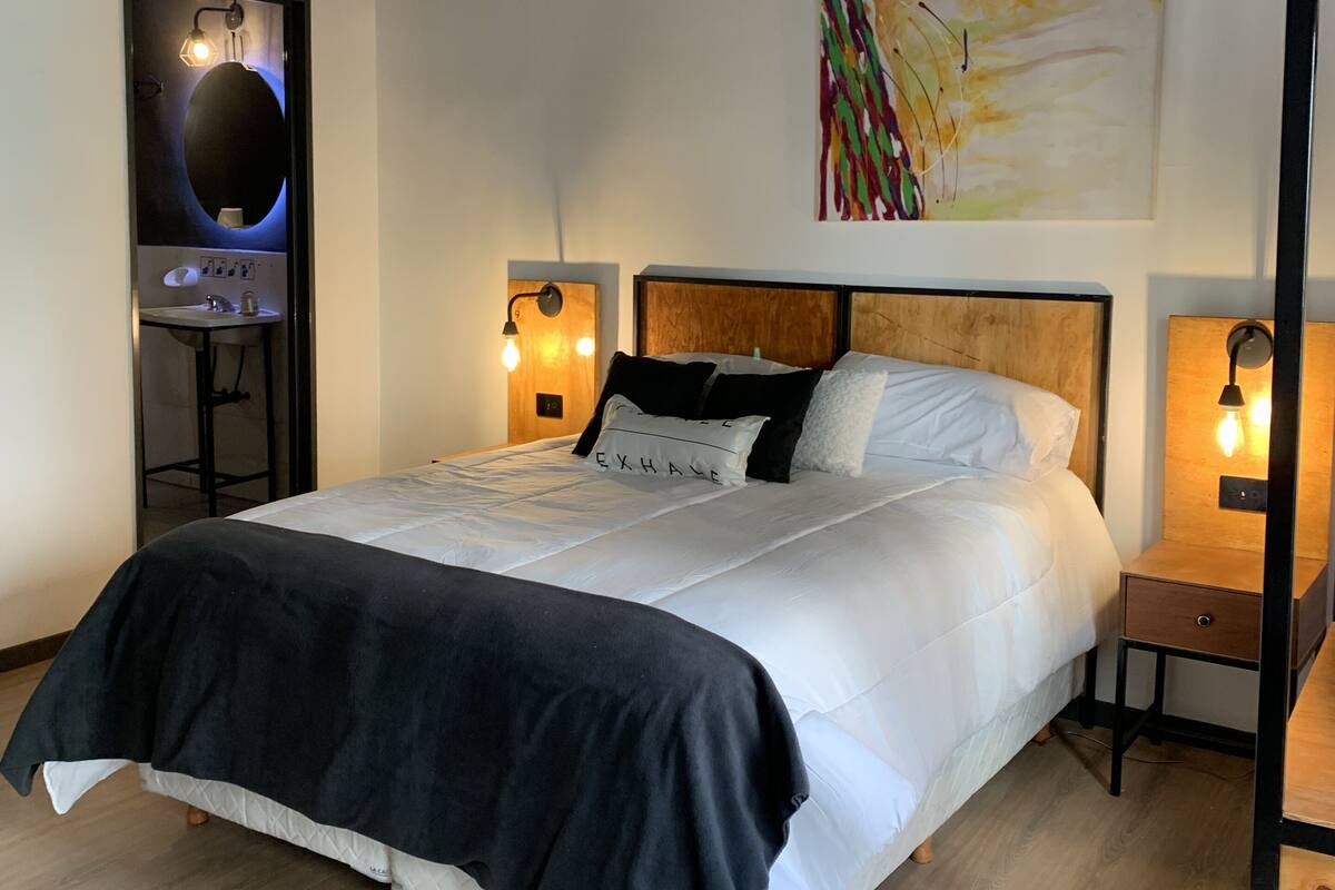 A neatly made bed with a black and white bedspread, two bedside tables with lamps, a wall-mounted abstract painting, and a mirror with a circular frame.