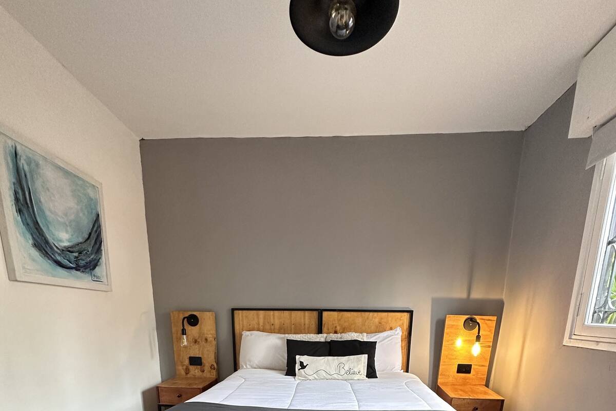 A bedroom with a large bed, wooden headboard, bedside tables with lamps, a painting on the wall, and a window with a view.