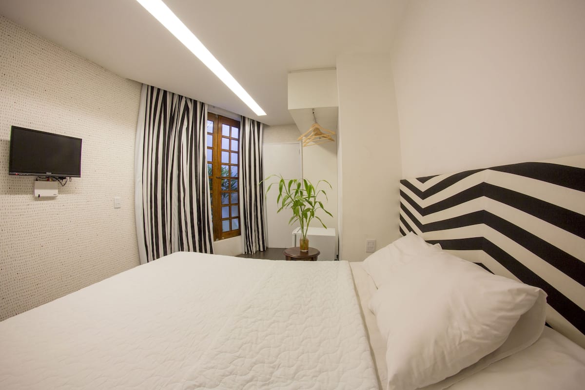 A neatly arranged hotel room with a bed, a television mounted on the wall, a window with striped curtains, and a hanging plant.
