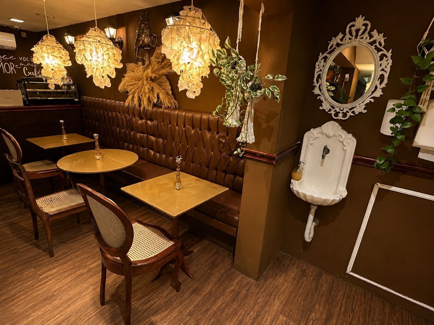 Un interior de restaurante con asientos de cuero marrón estilo cabina, mesas redondas con sillas, lámparas florales colgantes, un lavamanos blanco y un espejo decorativo.