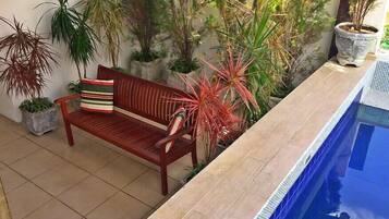 Uma área à beira da piscina com um banco de madeira, plantas em vasos e piso ladrilhado.