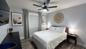Een slaapkamer met een bed, een plafondventilator, een nachtkastje met een lamp en kunstwerken aan de muur.