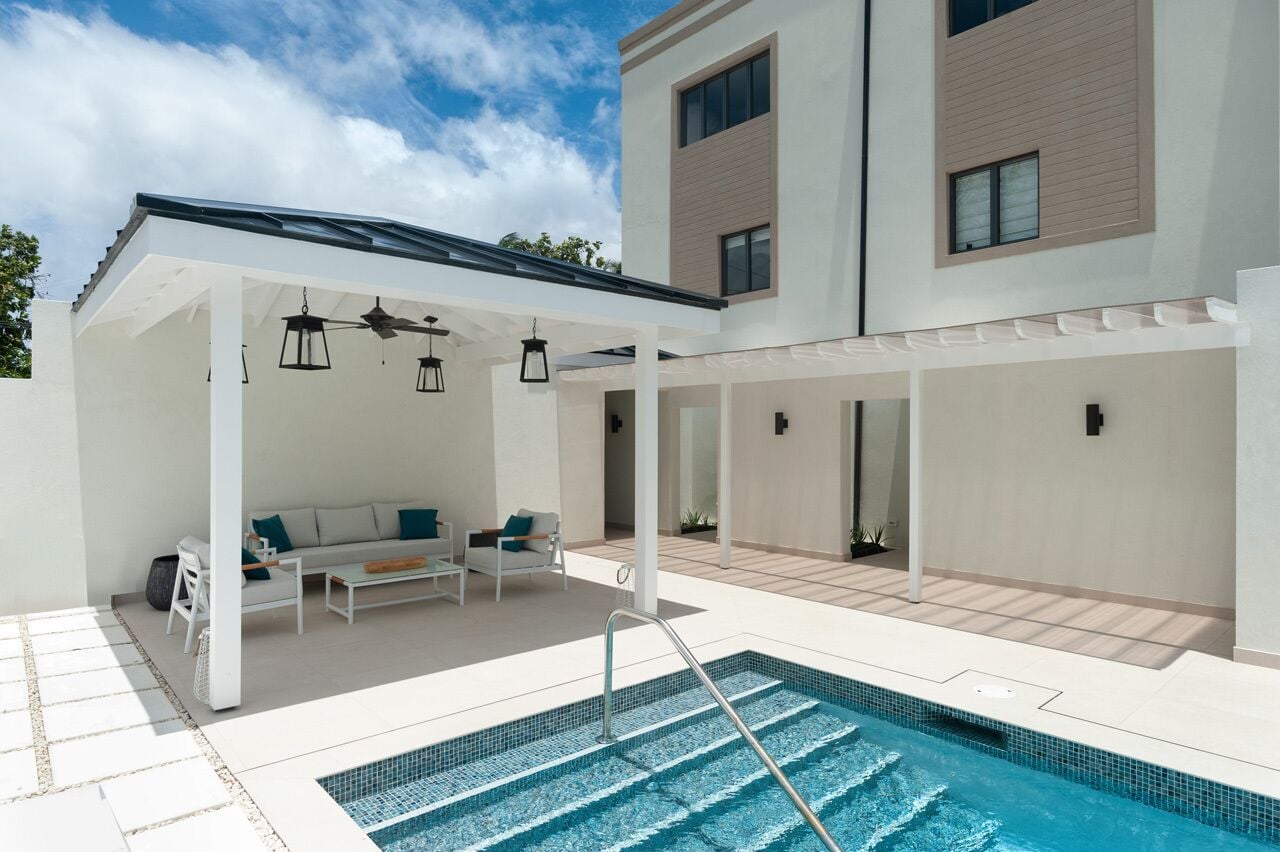 A modern outdoor pool area with a covered seating area, a pool with steps, and a building with a flat roof and large windows.
