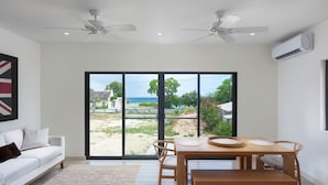 A modern living room with a white sofa, a wooden dining table with chairs, and a large sliding glass door leading to an outdoor area with a view of the sea. - Steps to Beach, Pool, Free Beach Club Access, Ocean/Sunset Views & Spacious! (Lower Carlton)