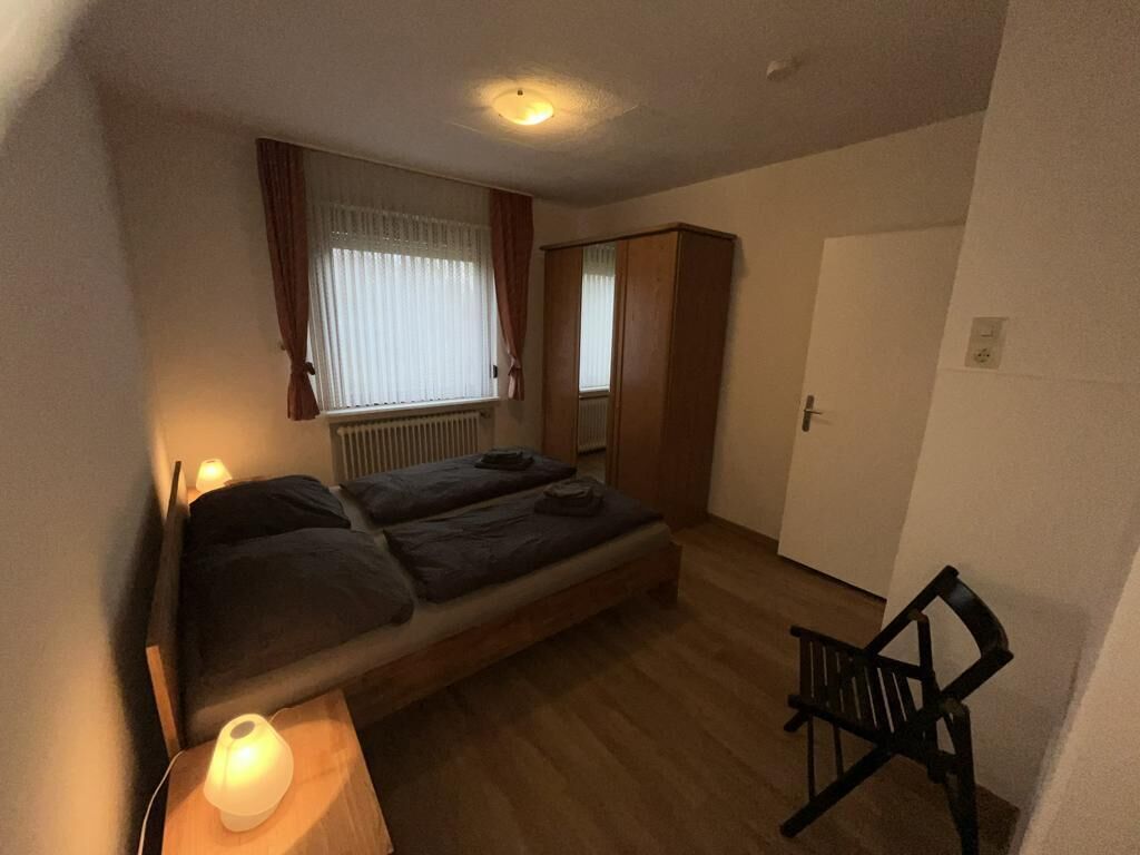 A small, single-room apartment with a bed, a nightstand, a chair, and a door.