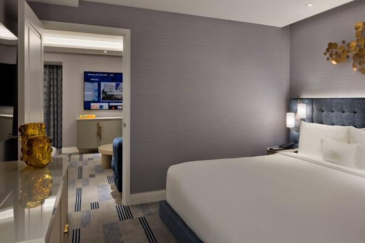 Une chambre d’hôtel moderne avec un grand lit, une tête de lit capitonnée, un élément décoratif mural et une petite table surmontée d’un vase.