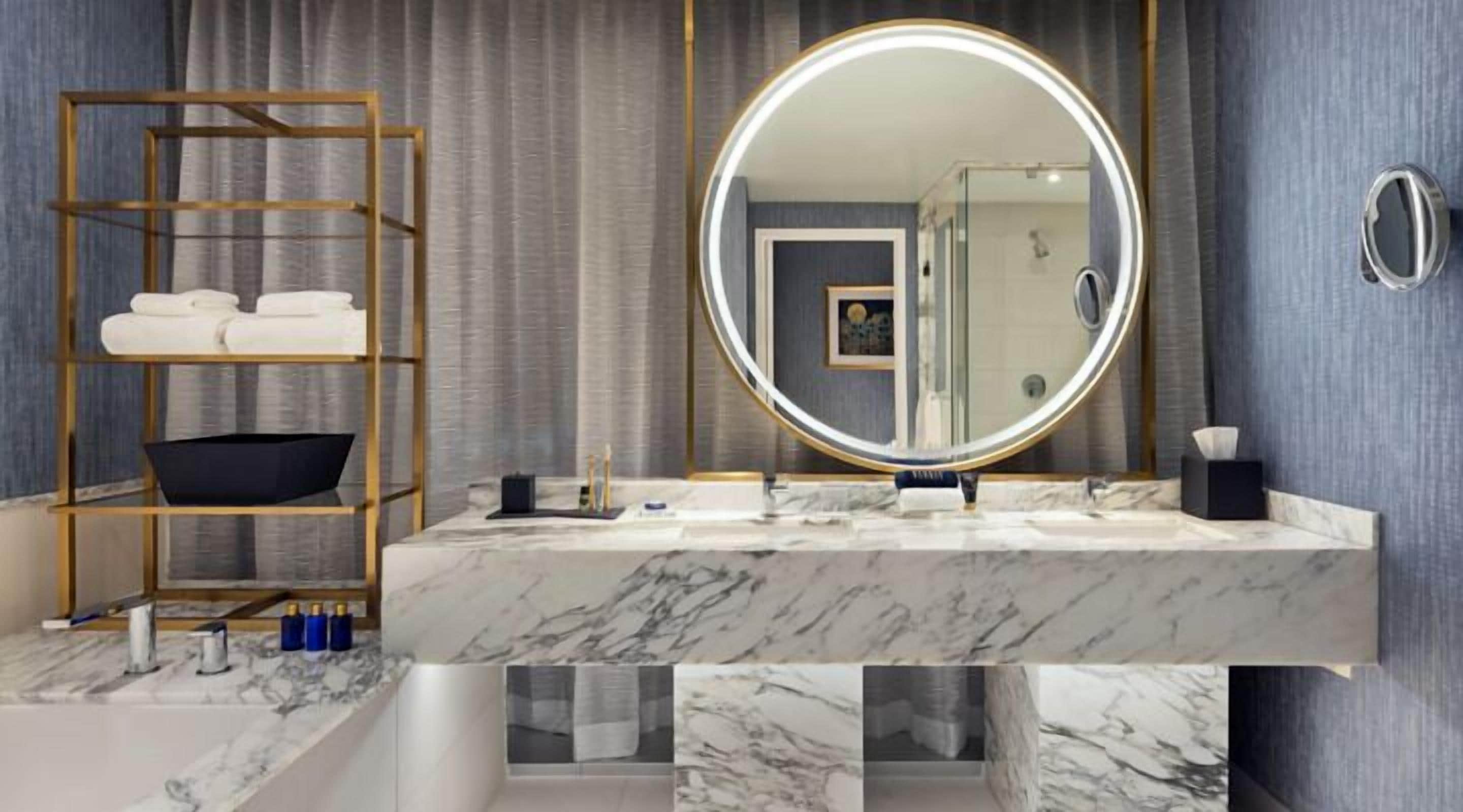 A bathroom with a marble vanity, a round mirror with a light, a wooden shelf with towels, and a glass shower door.