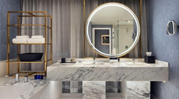 Un baño con tocador de mármol, un espejo redondo con luz, un estante de madera con toallas y una puerta de ducha de cristal.