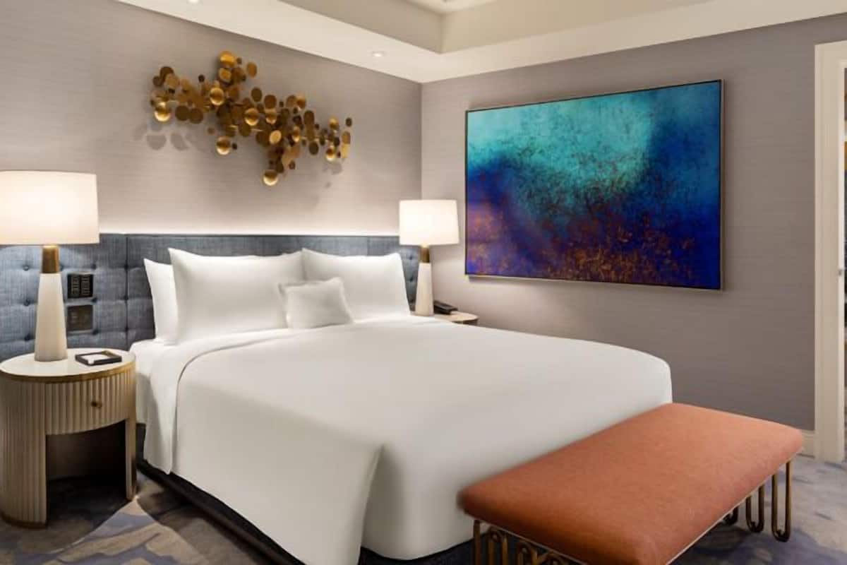 Une chambre d’hôtel moderne avec un grand lit, deux tables de chevet, un banc, une œuvre d’art murale et une grande fenêtre.