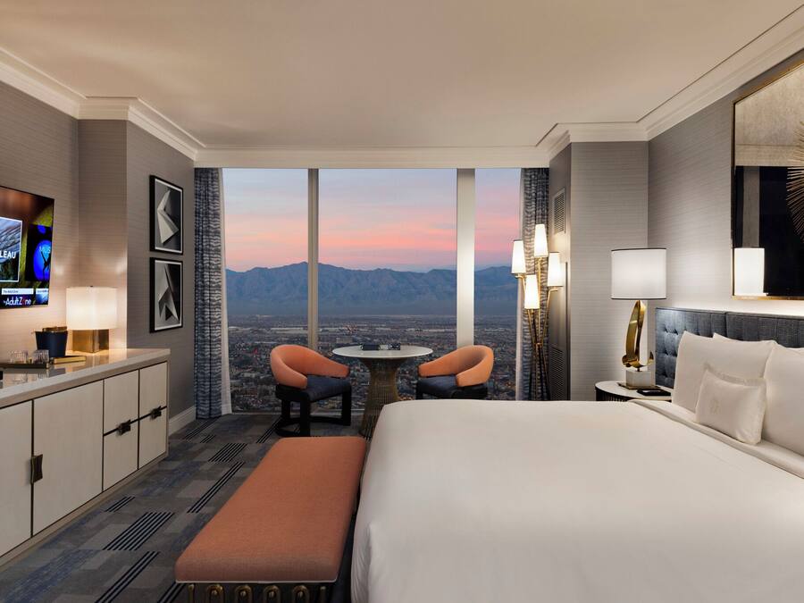 A hotel room with a large bed, a desk with a TV, two chairs, a small table, and a view of the mountains.