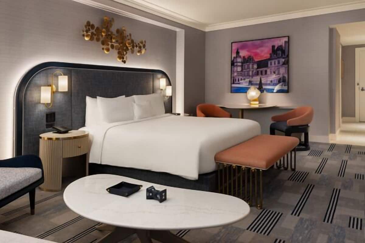 Une chambre d’hôtel moderne avec un grand lit, une table de chevet, un bureau et une chaise. Un tableau orne le mur et une fenêtre offre une vue sur les bâtiments.
