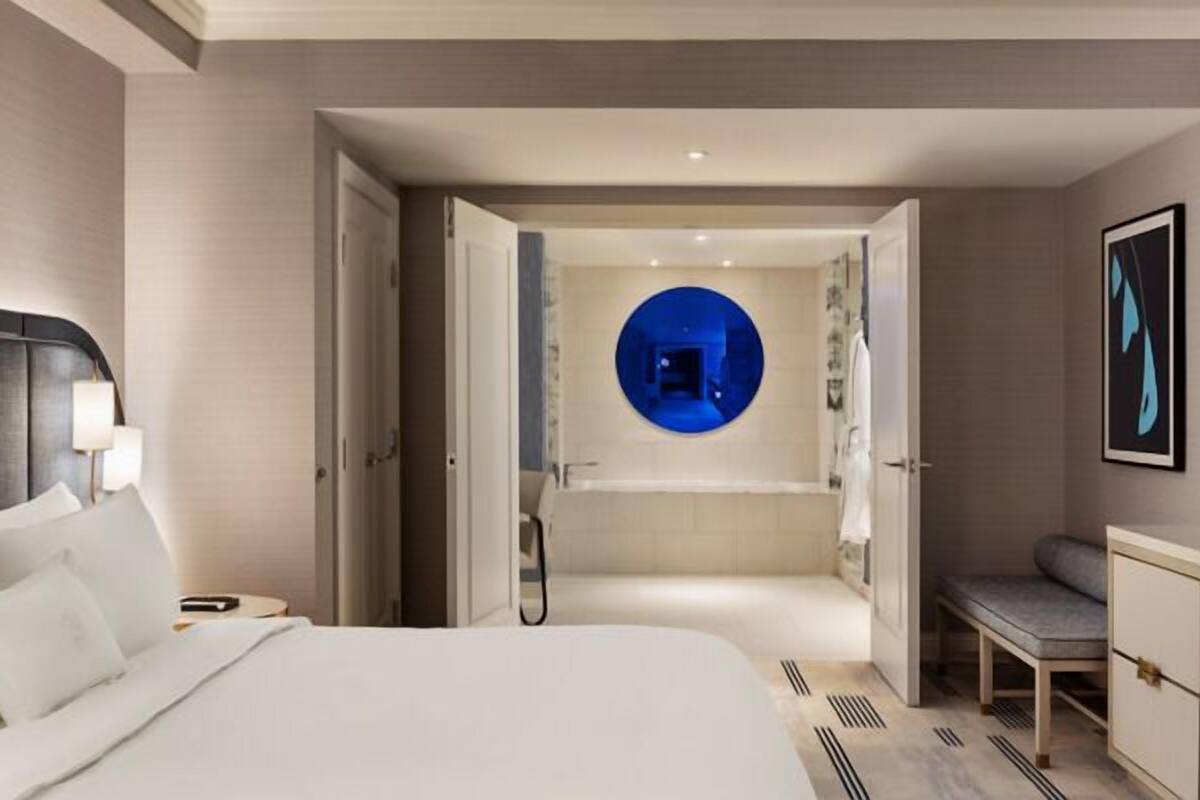 Une chambre d’hôtel moderne, dotée d’un grand lit, d’une table de chevet, d’un bureau et d’une salle de bain visible à travers une porte ouverte.